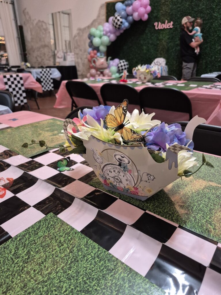 A tea party-themed table centerpiece shaped like a teapot, filled with colorful flowers and a decorative butterfly, sits on a grass-patterned and checkered tablecloth. Balloons and guests are in the background.