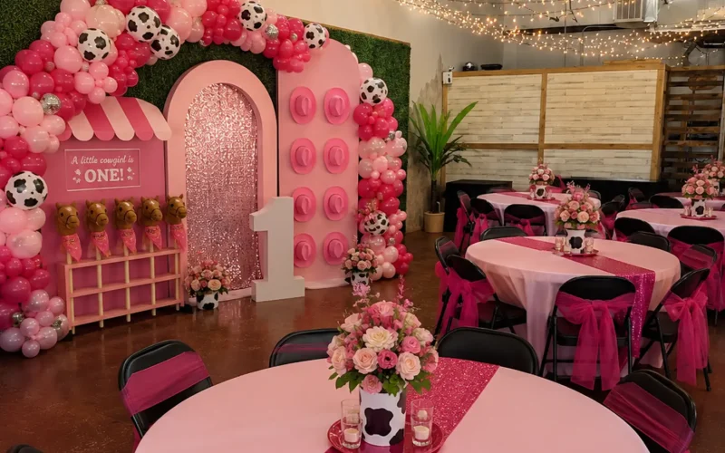 A farm-themed first birthday party with pink cow print balloons, a large “1,” pink tablecloths, flower centerpieces, and plush cows on display. String lights hang above decorated round tables with black chairs.